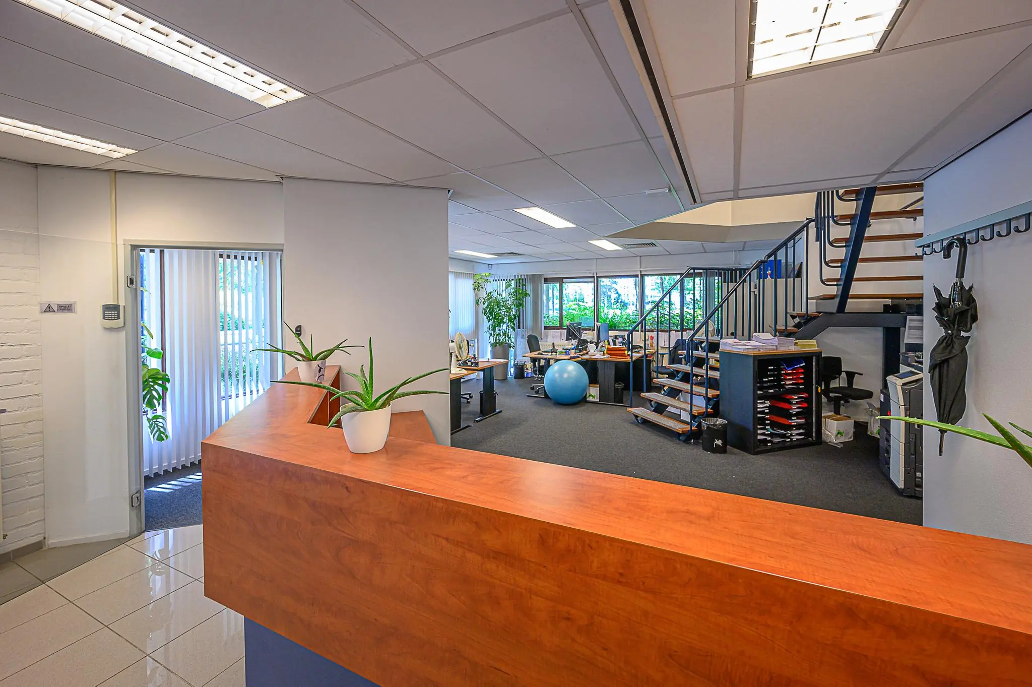 Moderne kantoorruimte met receptiebalie, bureaus, een trap naar boven en veel natuurlijk licht.